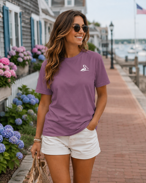 A woman wearing a berry colored short-sleeve t-shirt with a small white Beak & Tide pelican logo on the left chest, paired with white shorts, walking along a brick boardwalk next to flowers and water.